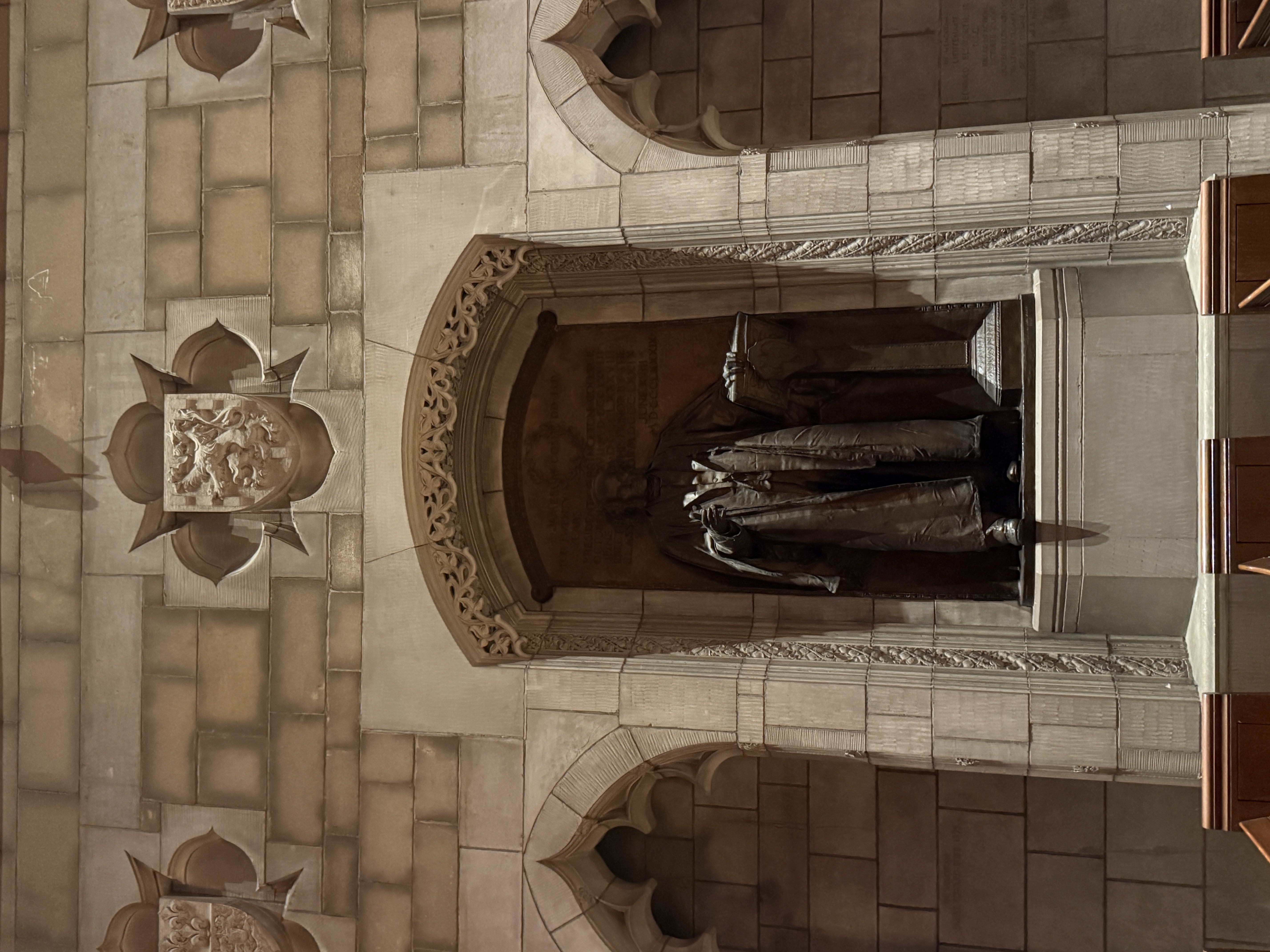 Statue of a robed figure standing in a recessed niche beneath heraldic shields
