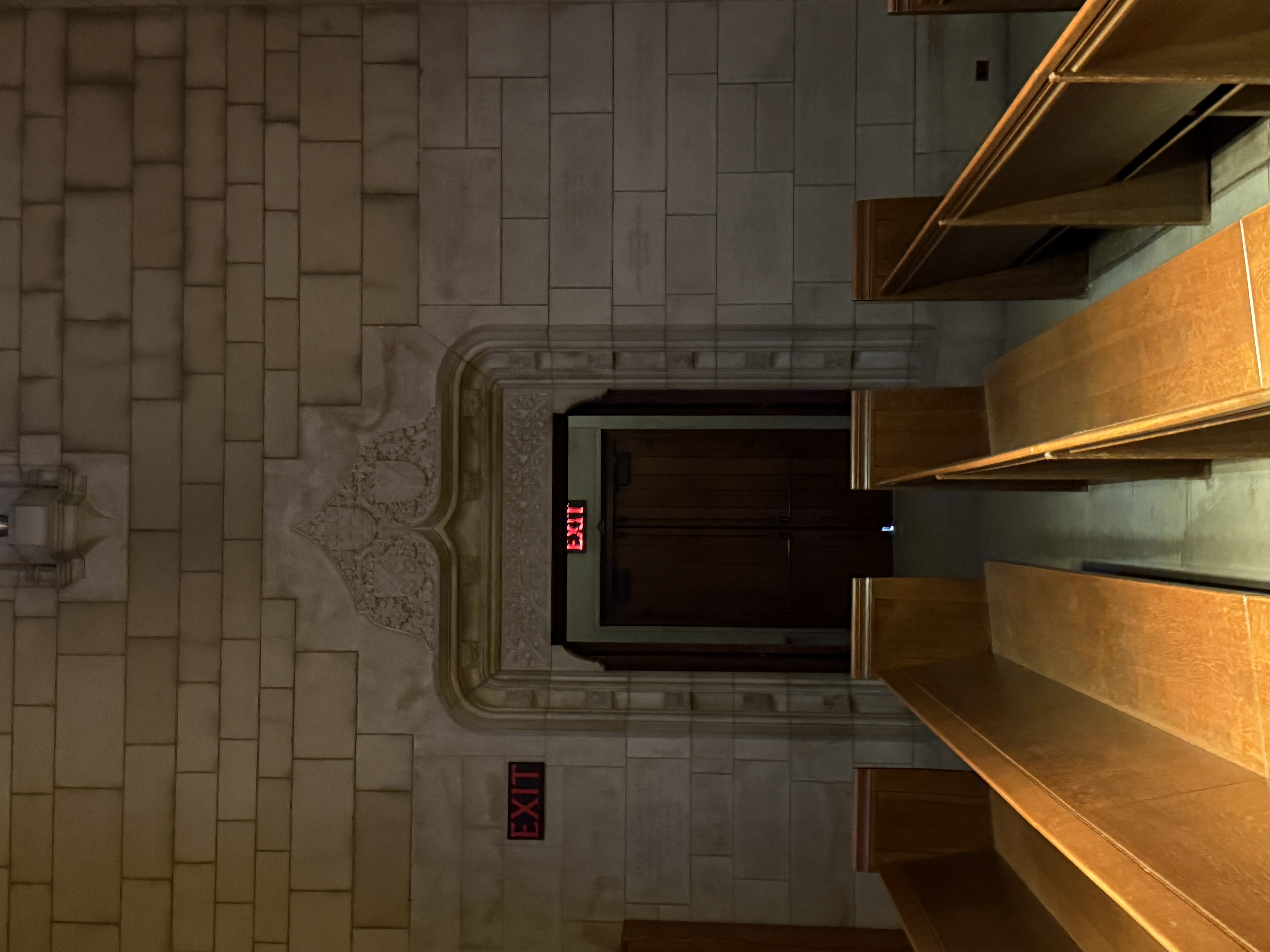 Interior aisle between wooden pews facing a doorway with red exit signs